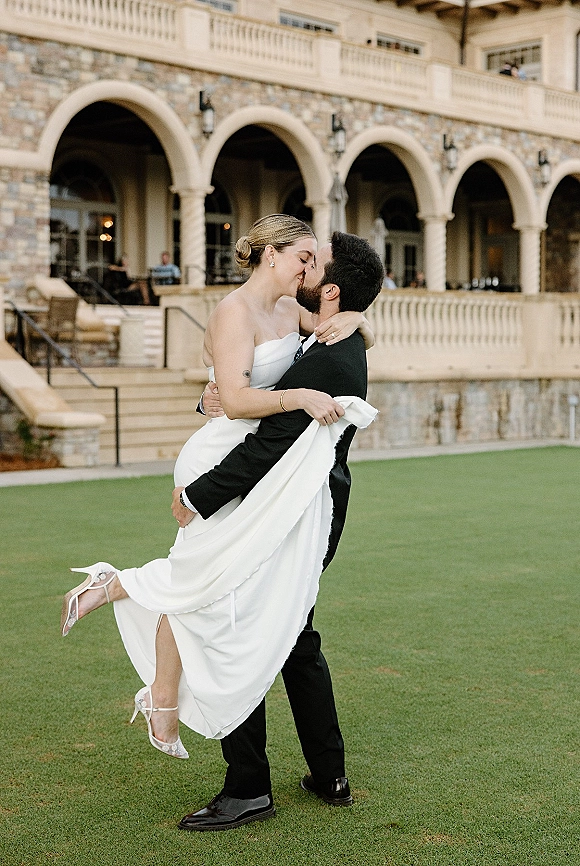 Wedding kiss portrait of groom lifting bride in a strapless dress as they kiss before stone arches and a grand staircase backdrop
