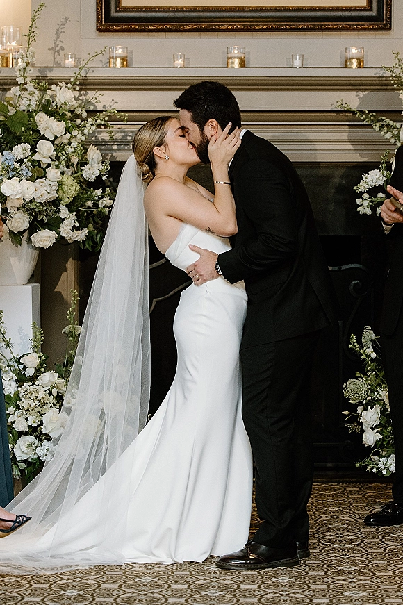 Wedding kiss as bride in strapless gown and cathedral veil embraces groom in tux by a candlelit fireplace mantel with florals