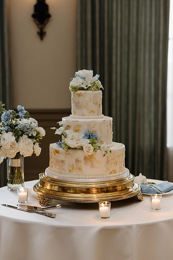 Wedding cake with textured buttercream and fresh flowers on a gold stand, set on a candlelit table in an indoor reception room