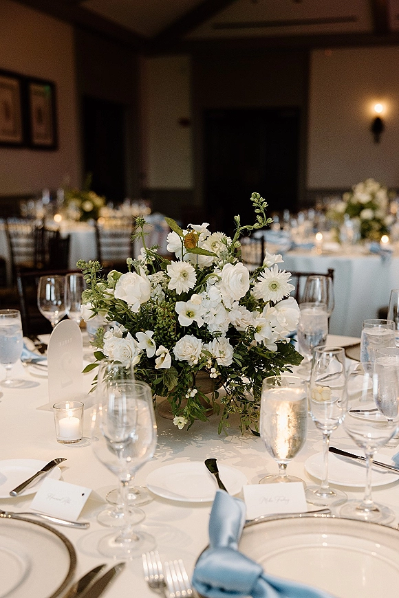 Reception tablescape with a white floral centerpiece, blue napkins, place cards, and votive candles on round tables in an indoor reception room