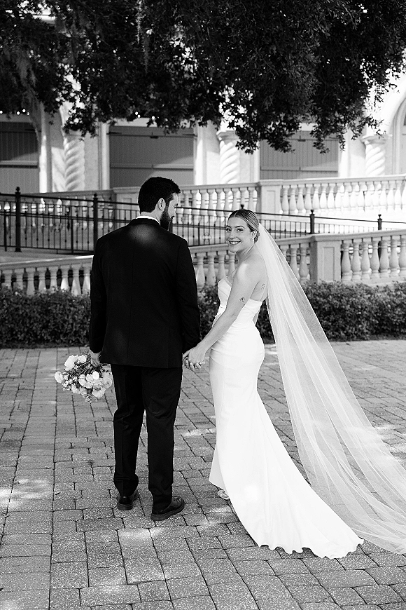 Couple portrait in black and white, bride and groom holding hands as they walk away in a stone courtyard with trees and columns, long veil trailing
