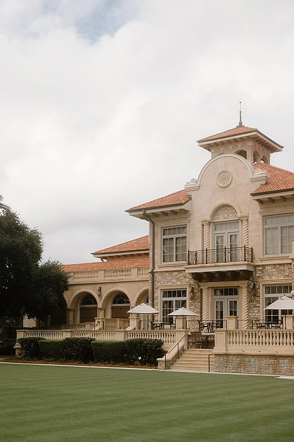 Wedding venue exterior with stone facade and arched colonnade, balcony with wrought iron railing, terrace tables on lawn under cloudy sky