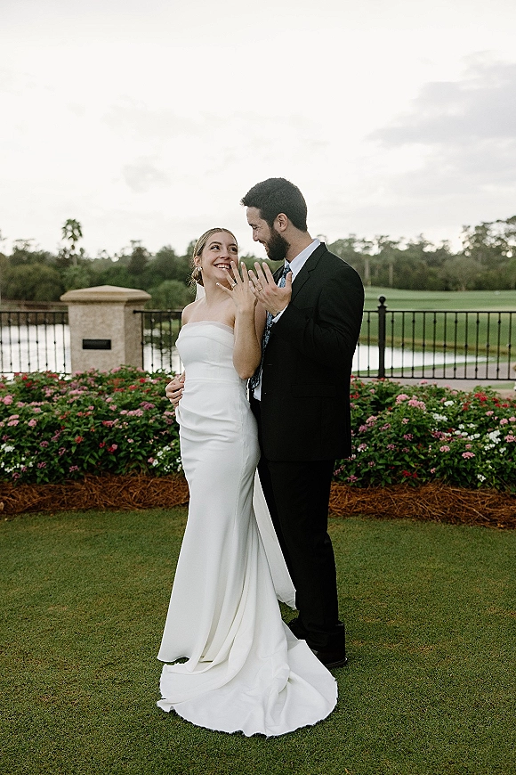Couple portrait of bride and groom hugging, bride showing ring with veil and strapless dress on a garden lawn by a lake under cloudy sky