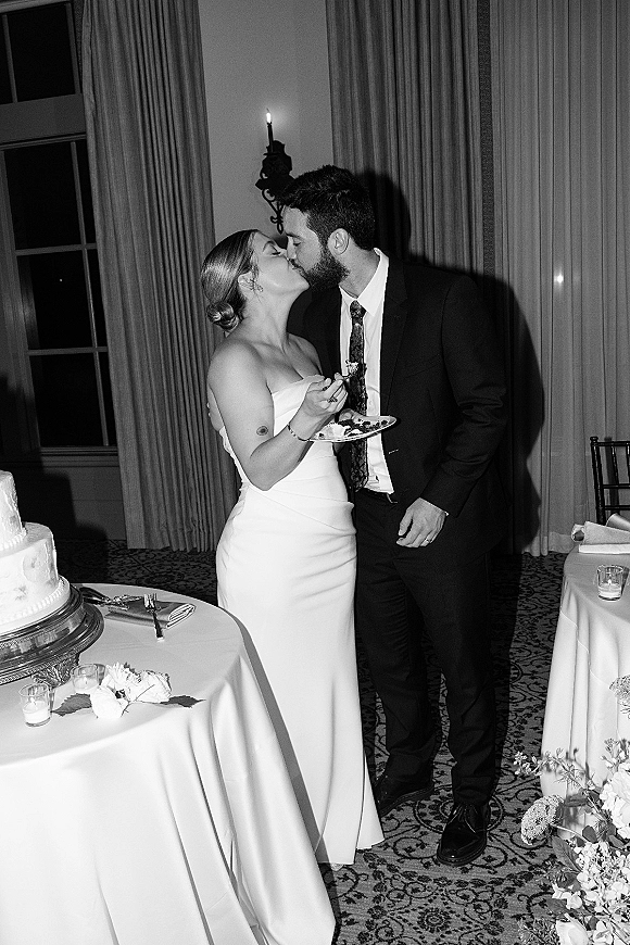 Wedding cake cutting as bride in strapless gown feeds groom cake beside tiered cake with votive candles and white florals, draped curtains behind