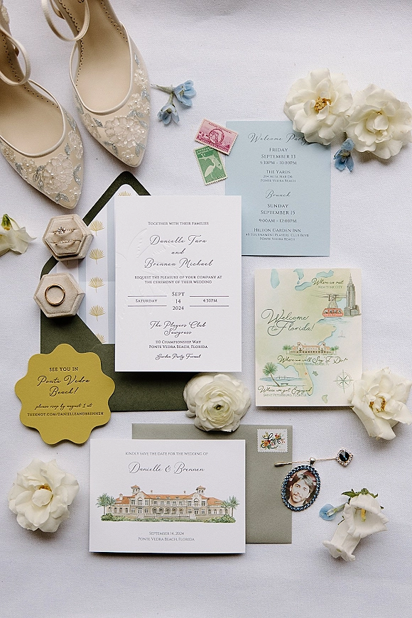 Wedding invitation suite flat lay with envelopes, RSVP and map cards, stamps, ring box, bridal shoes, and flowers on white linen fabric