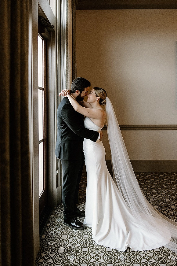 Wedding couple portrait of bride and groom embrace in soft window light, long veil trailing as he gives a forehead kiss in a hotel hallway