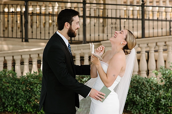 Wedding vows as bride reads from a vow booklet in wedding dress and veil while groom in suit listens by an iron fence and greenery hedge