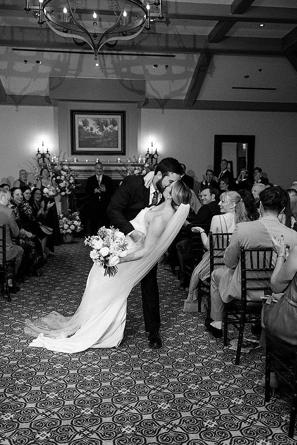 Wedding kiss as groom dips bride in strapless dress and veil, bouquet in hand, with cheering guests in candlelit indoor aisle