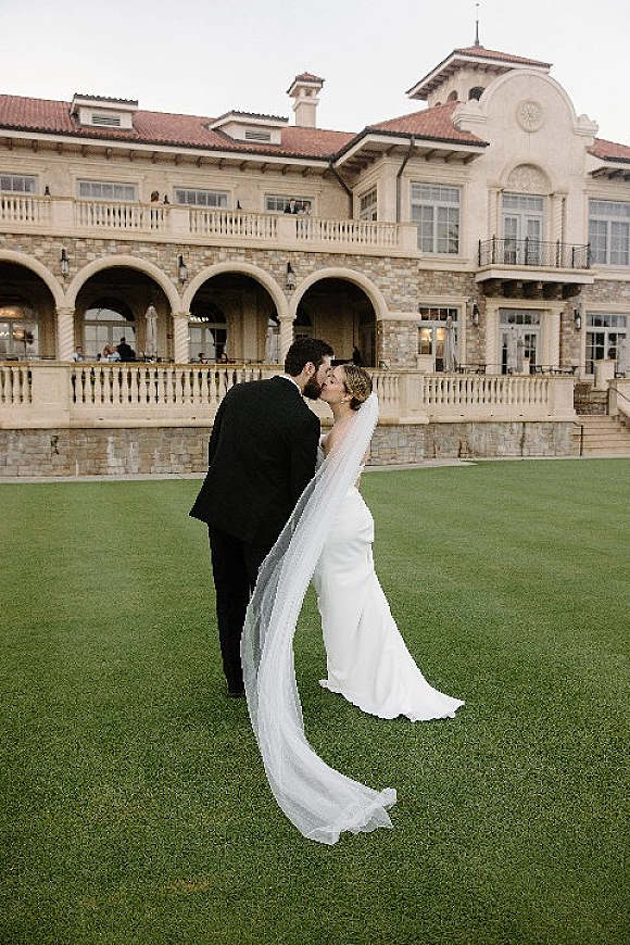 Wedding kiss as bride and groom embrace on a manicured lawn, her long bridal veil trailing behind, with a stone villa and arches backdrop