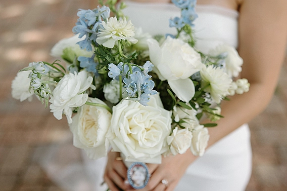 Bridal bouquet of white roses and blue delphinium with greenery, held against a bridal gown, with engagement ring visible in natural light