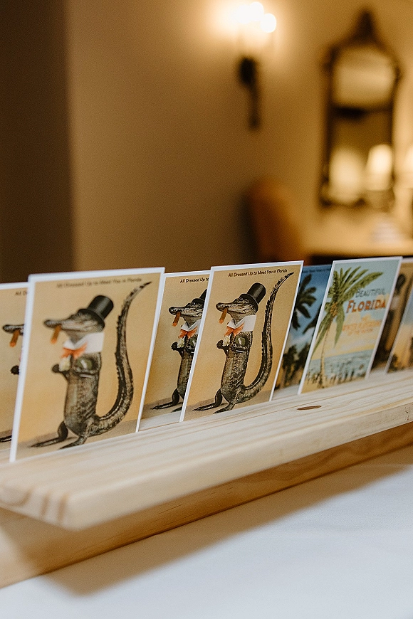 Wedding escort cards arranged in a wooden holder, illustrated postcard-style designs on a ledge beneath a wall sconce and mirror indoors