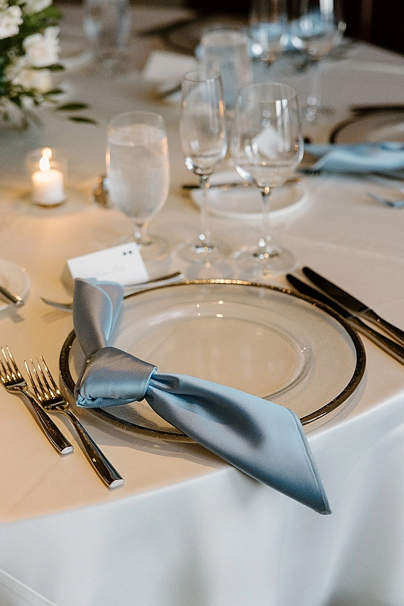 Reception tablescape with wedding place setting, light blue napkin knot, gold rim charger, candles, and florals on a softly lit banquet table