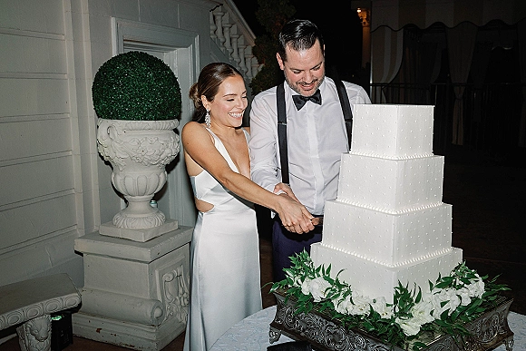 Wedding cake cutting with bride and groom cutting cake beside a four-tier white cake on a silver stand, framed by greenery garland on a patio at night