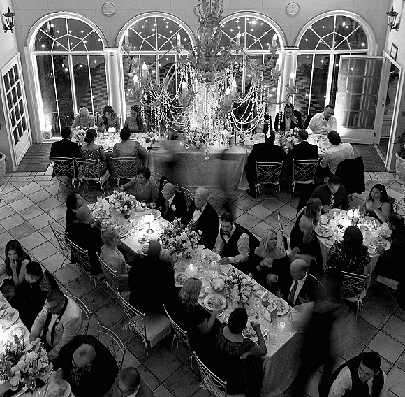 Wedding reception with guests dining beneath a crystal chandelier, candlelit tables with floral centerpieces in an arched-window hall