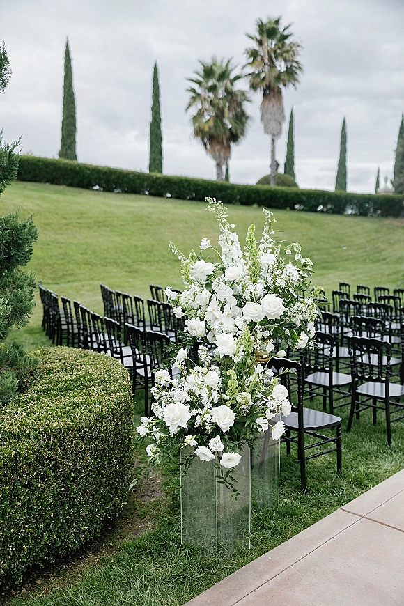 Ceremony aisle decor with a white floral arrangement and greenery on a clear acrylic pedestal beside black Chiavari chairs on a lawn