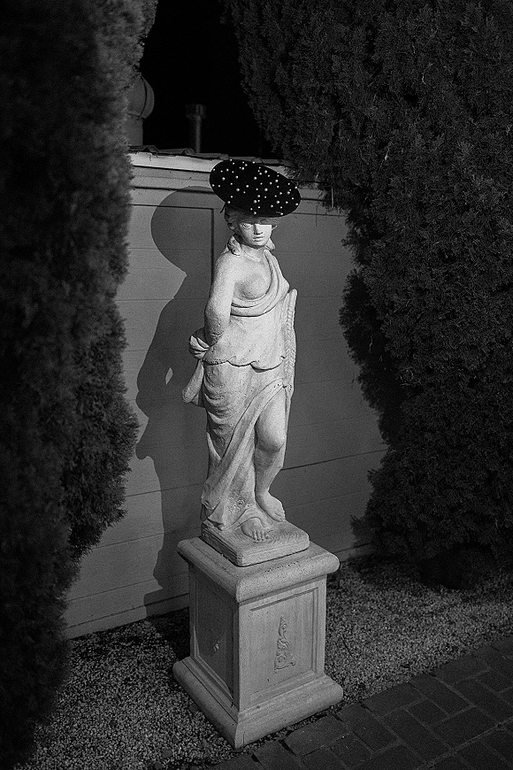 Garden statue, stone garden statue of a classical woman on a pedestal with wide brim hat, set between hedges by a garden wall and gravel path