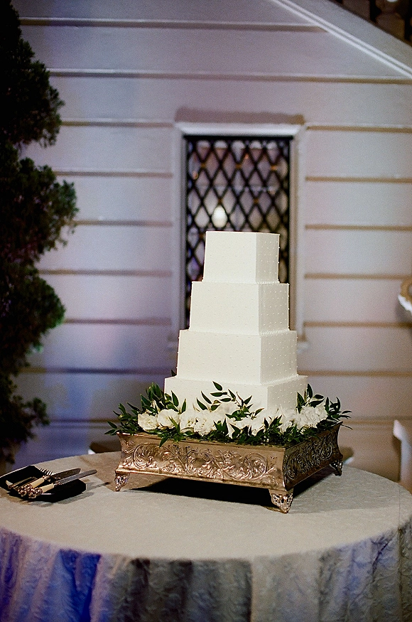 Wedding cake with a square tier wedding cake design, embossed white buttercream on ornate silver stand, greenery garland and flowers on round table outdoors