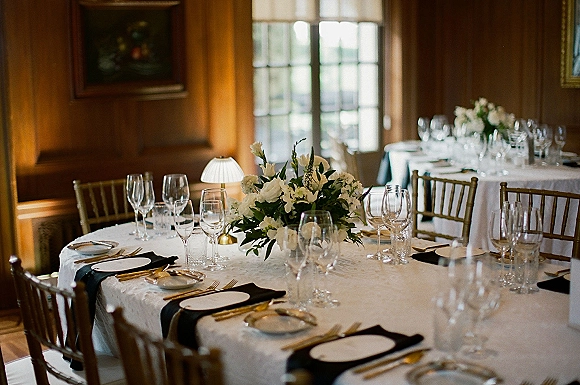 Reception tablescape with round wedding table setup, white floral centerpiece with greenery, black napkins, gold flatware, and candles in a wood-paneled room