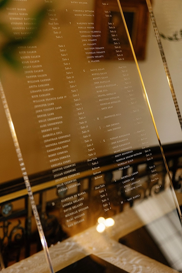 Wedding seating chart mirror seating chart in a gold frame on an easel, displaying printed guest list in a warm-lit reception space