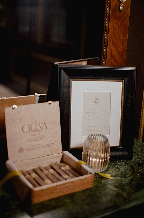 Wedding welcome table with a framed sign and open cigar box display beside a ribbed glass votive candle holder on green marble in a dim interior