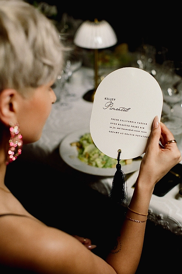 Wedding menu card with calligraphy wedding menu text and a black tassel, styled beside a plate and glassware on a reception table setting