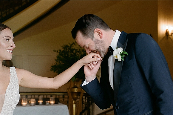 Wedding couple portrait of groom kissing bride hand as they hold hands on an indoor staircase, bride in strapless lace dress with orchid boutonniere