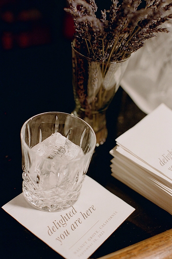 Wedding place cards with calligraphy place cards beside a rocks glass with ice and dried florals in a vase on a dark tabletop