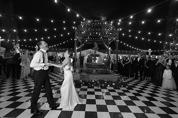 First dance as bride in satin gown and groom in suspenders hold hands on a checkered floor under string lights in a courtyard at night