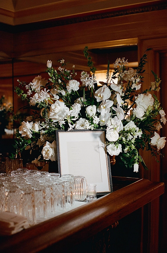 Wedding welcome table with a framed sign, white floral arrangement and greenery, glassware and votive candles on a wood-paneled bar counter