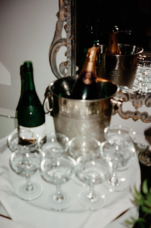 Champagne bar setup with chilled bottles in ice buckets, wine glasses on a bar cart, and an ornate mirror against a white wall