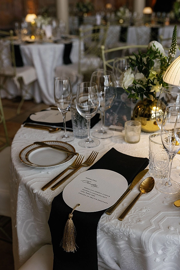 Reception tablescape with a black and gold wedding table setting on a white textured tablecloth, candlelight and floral centerpiece in warm banquet room