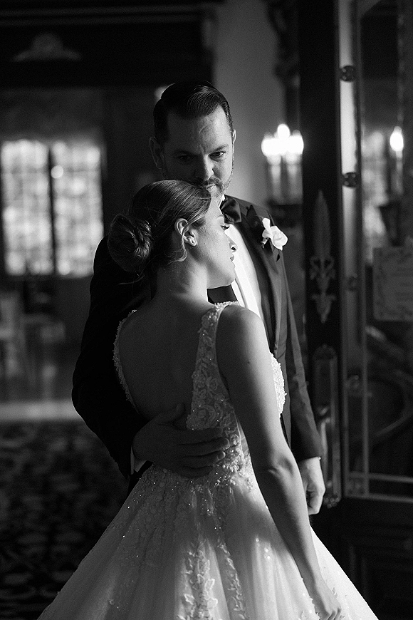 Couple portrait of bride and groom embrace in a doorway, lace gown and black tuxedo with rose boutonniere in window light