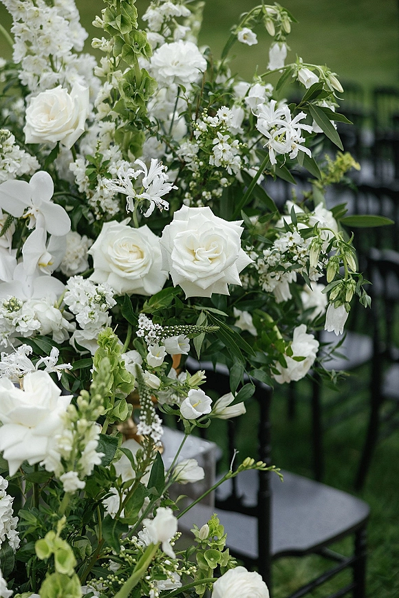 Wedding floral arrangement of white wedding flowers with roses and orchids spilling over greenery on black ceremony chairs on an outdoor lawn