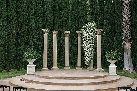 Ceremony backdrop with white floral arrangement and greenery on stone columns, set before a tall evergreen hedge on a lawn