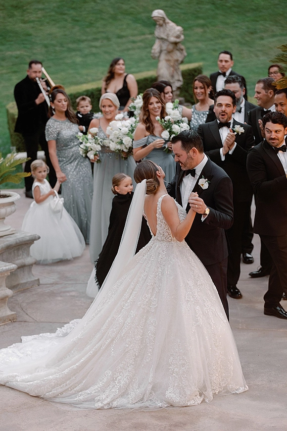 Wedding recessional as bride and groom walk out arm in arm, long lace train and veil flowing as wedding party cheers on a stone terrace