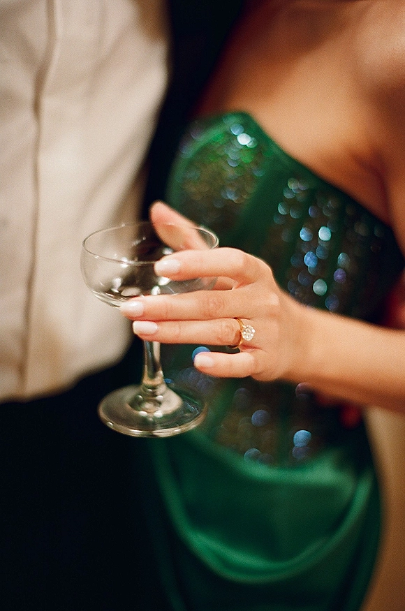 Engagement ring on hand beside a wine glass, diamond ring catching light over a green sequin dress, with a blurred couple behind