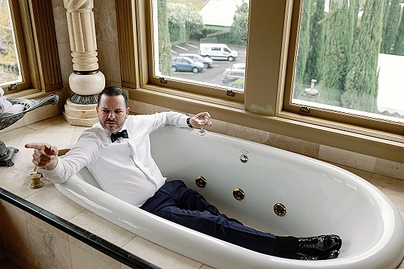 Groom portrait with groom getting ready photo vibe, wearing a tuxedo bow tie in a bathtub, holding a champagne glass by large windows