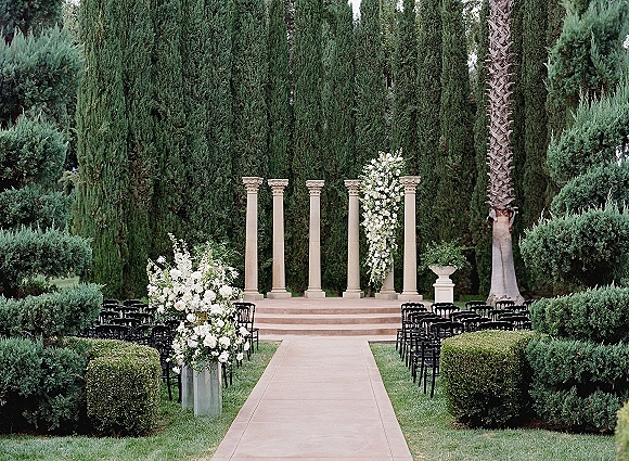 Ceremony setup with an outdoor ceremony aisle lined by black chairs, white floral urns, and stone columns on a manicured lawn with cypress trees