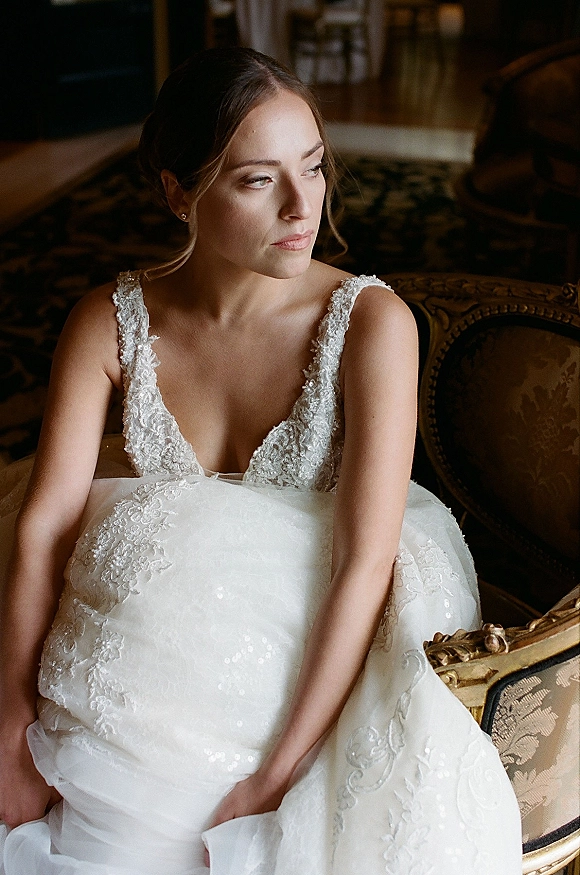 Bridal portrait of a bride sitting on an upholstered armchair in a lace wedding dress, looking away in soft window light indoors