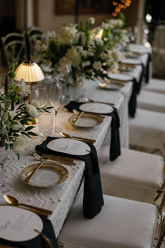 Reception tablescape on a long banquet table with white floral centerpiece and greenery, black napkins, gold flatware, under warm string lights