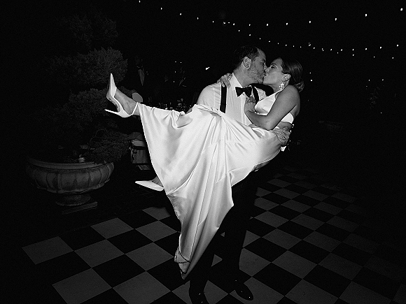 Wedding kiss portrait of groom carrying bride as they kiss, her satin gown and statement earrings glowing under string lights on a patio at night