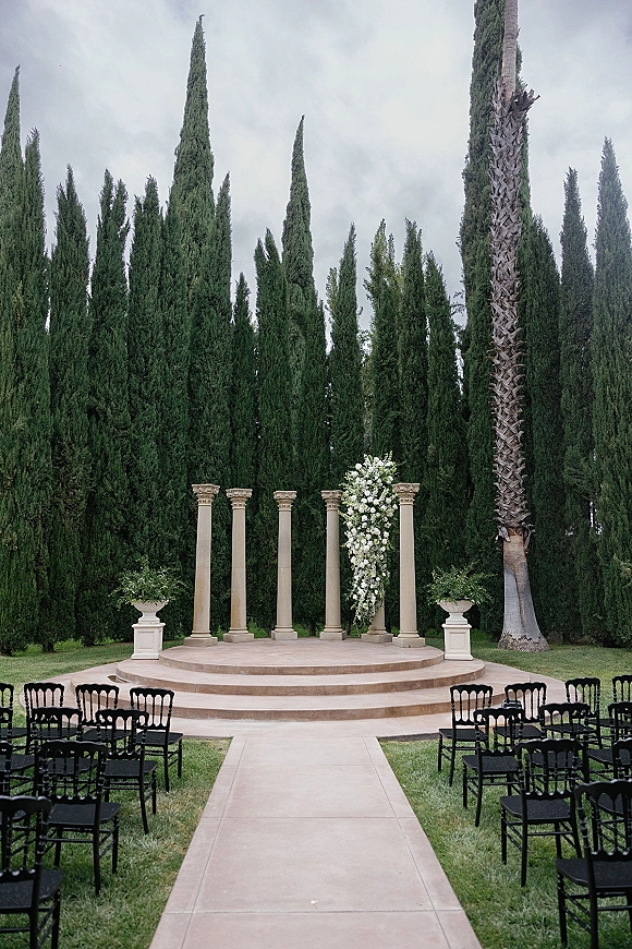 Outdoor ceremony setup with a white floral arrangement on a round stage between stone columns, black chairs lining an aisle on lawn