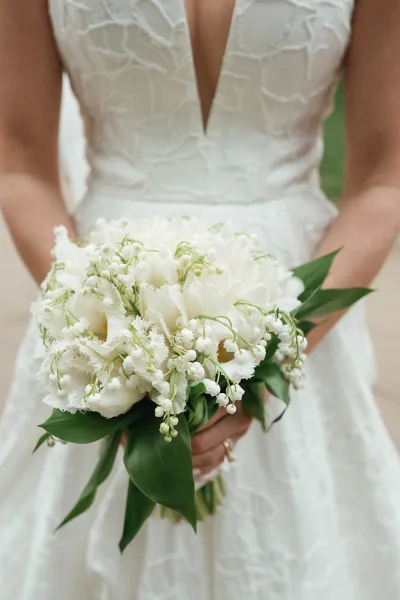 Bridal bouquet of white tulips and lily of the valley held against a lace deep V gown, with green foliage and soft blurred greenery behind