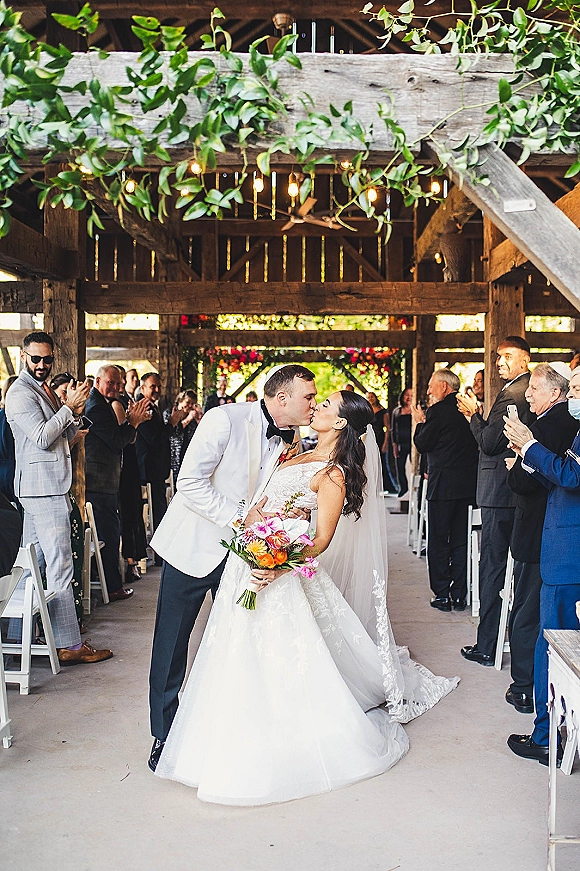 Wedding kiss as bride holds bouquet and leans into groom in tuxedo, guests cheering in a rustic barn aisle with string lights