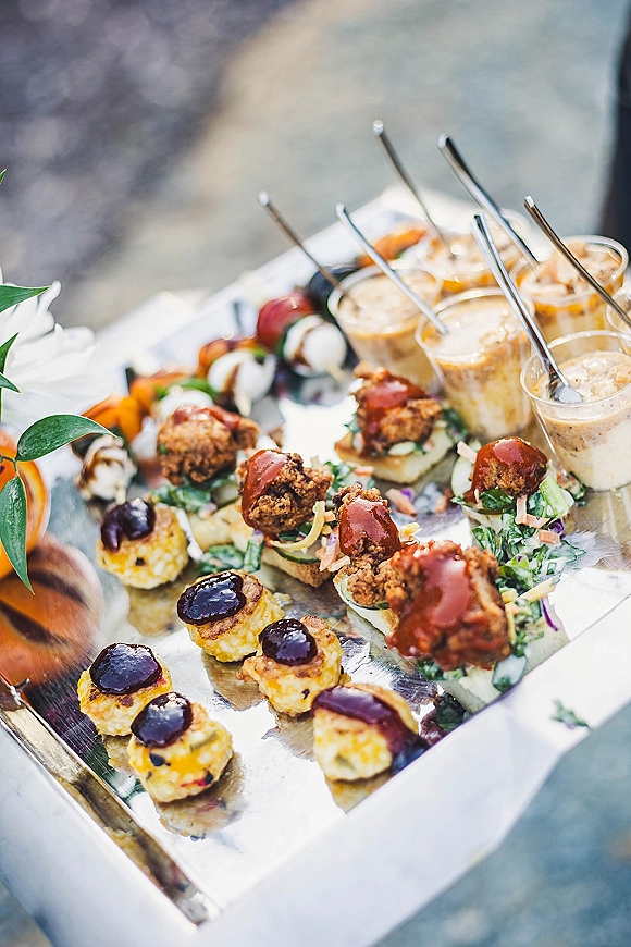 Wedding appetizers on a silver tray with cocktail hour food in skewers and mini cups, sauce drizzle and greenery on a stone surface outdoors