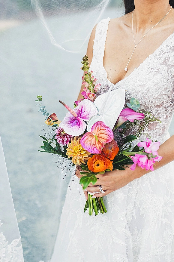 Bridal bouquet with tropical wedding bouquet blooms and greenery held against a lace wedding dress, with veil and pearl necklace on a soft neutral backdrop