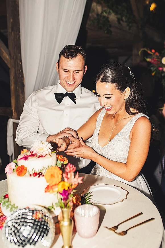 Wedding cake cutting as bride in a bridal gown and groom in a black bow tie slice a two-tier cake with colorful flowers under warm drapery