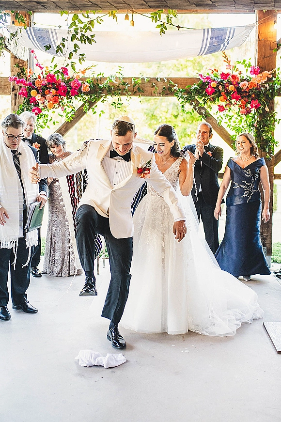 Wedding recessional at a Jewish wedding ceremony as bride in lace veil and groom in white tux jacket walk under a floral chuppah on a lawn