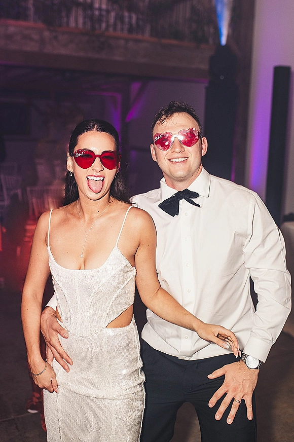 Reception party portrait of a couple in heart-shaped sunglasses, bride in sequin dress and groom in bow tie on a hazy dance floor
