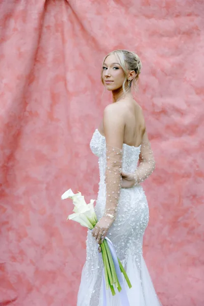 Bridal portrait of a bride holding bouquet in a strapless lace wedding dress, sheer pearl gloves, against a pink textured wall backdrop